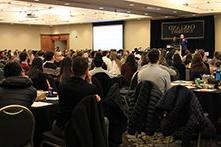 A large room full of people seated at tables, listening to a presentation.